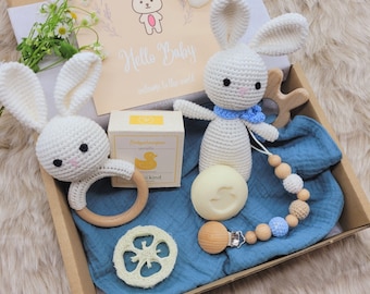 May include: A gift box filled with baby items, including two crocheted white bunny toys, a wooden teether, a blue and white beaded necklace, a bar of soap, a loofah, and a card that says "Hello Baby, welcome to the world".