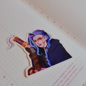 May include: A sticker of a character with blue hair and a dark blue cloak, holding a weapon, is placed on an open notebook. The notebook has a grid pattern and the date "APR 11" is visible. The background is a soft pink.