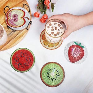 May include: A selection of fruit-shaped coasters, featuring a watermelon, kiwi, orange segment, and strawberry. The coasters are decorated with rhinestones. An apple coaster is in a black metal stand.