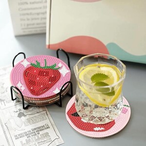 May include: A set of strawberry-themed coasters with a black metal holder. Each coaster features a pink background with a red strawberry design. A glass of water with lemon slices and mint sits on a matching coaster.