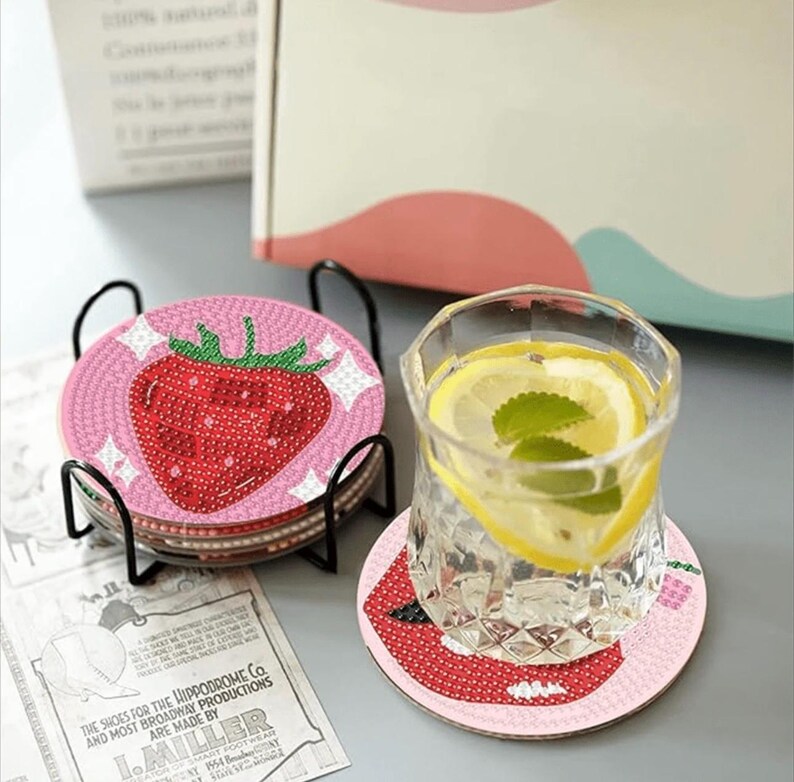 May include: A set of strawberry-themed coasters with a black metal holder. Each coaster features a pink background with a red strawberry design. A glass of water with lemon slices and mint sits on a matching coaster.