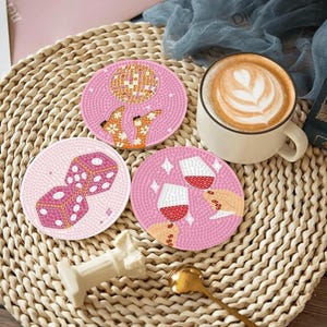 May include: Set of three pink coasters with embroidered designs: dice, wine glasses clinking, and a disco ball with legs. A cup of coffee with latte art sits nearby. The coasters are on a woven mat.
