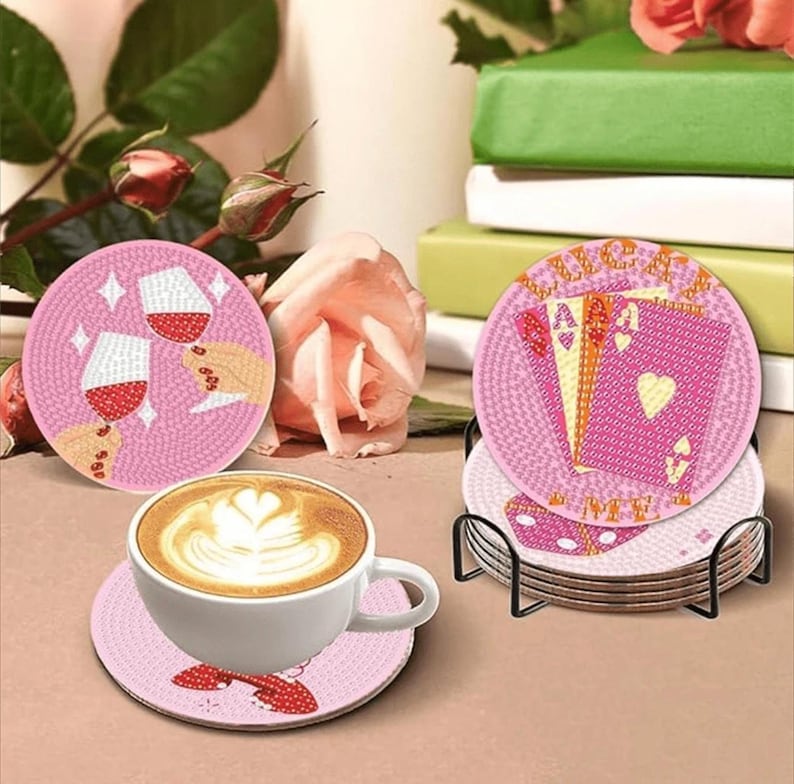 May include: Pink coasters with various designs, including wine glasses, playing cards, and the text "ME". A white coffee cup with latte art sits on a matching coaster. The coasters are displayed with a black metal holder.