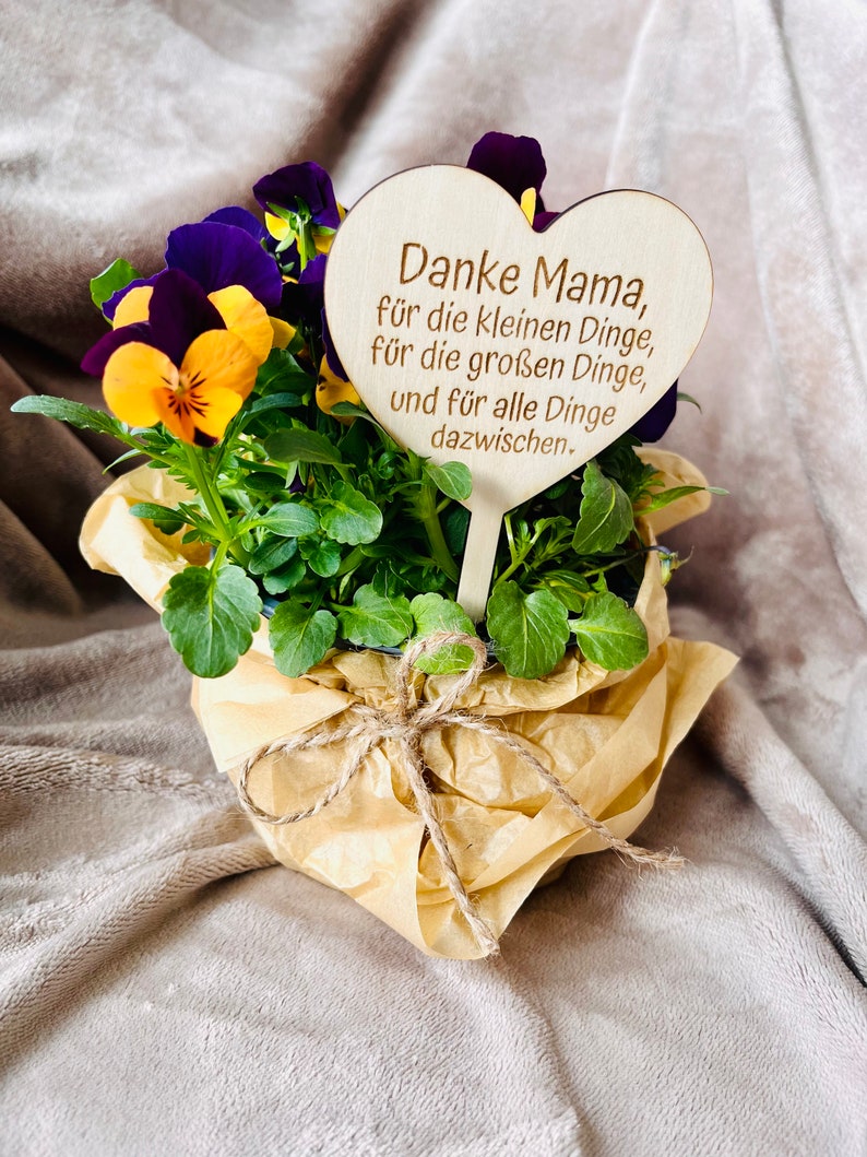 May include: A small potted plant with purple and yellow flowers, wrapped in brown paper and tied with twine. A wooden heart-shaped sign with the text "Danke Mama, f&uuml;r die kleinen Dinge, f&uuml;r die gro&szlig;en Dinge, und f&uuml;r alle Dinge dazwischen." is attached to the plant.