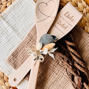 May include: Two wooden spoons tied together with twine and dried flowers. One spoon has a heart carved into it and the other has the text "Liebe ist die wichtigste Zutat im Rezept des Lebens" which translates to "Love is the most important ingredient in the recipe of life."
