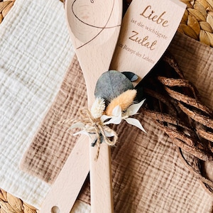 May include: Two wooden spoons tied together with twine and a small bouquet of dried flowers. One spoon has a heart carved into it, and the other has the text "Liebe ist die wichtigste Zutat im Rezept des Lebens" which translates to "Love is the most important ingredient in the recipe of life."