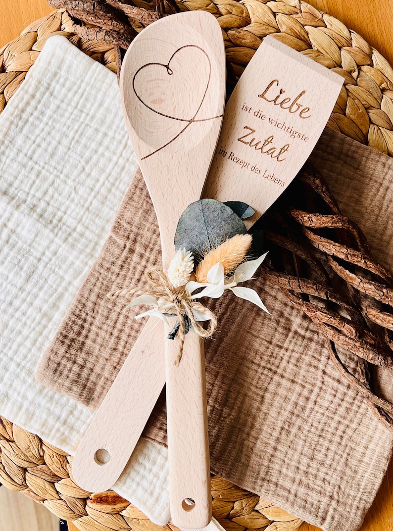 May include: Two wooden spoons tied together with twine and a small bouquet of dried flowers. One spoon has a heart carved into it, and the other has the text "Liebe ist die wichtigste Zutat im Rezept des Lebens" which translates to "Love is the most important ingredient in the recipe of life."