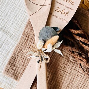 May include: Two wooden spoons tied together with twine and dried flowers. One spoon has a heart carved into it, the other has the text "Liebe ist die wichtigste Zutat im Rezept des Lebens".
