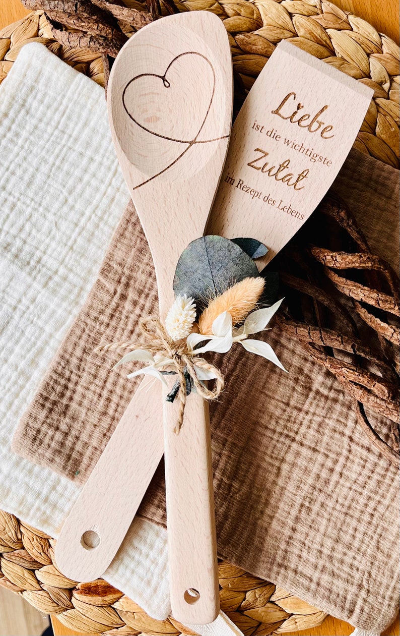 May include: Two wooden spoons tied together with twine and dried flowers. One spoon has a heart carved into it, the other has the text "Liebe ist die wichtigste Zutat im Rezept des Lebens".