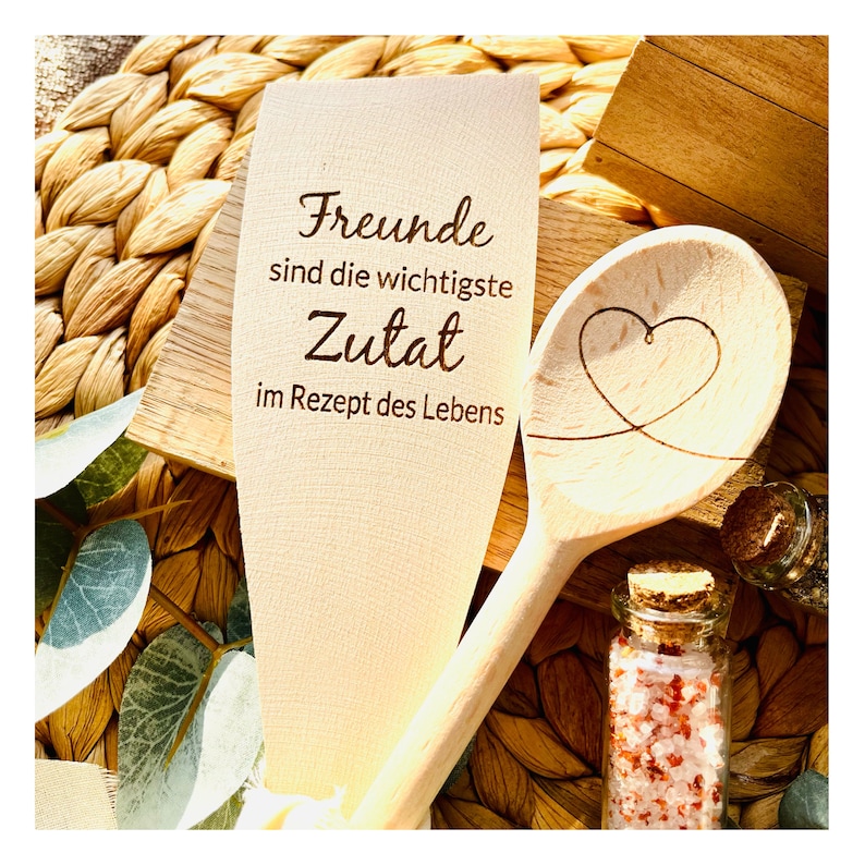 May include: Wooden spoon with the text "Freunde sind die wichtigste Zutat im Rezept des Lebens" and a heart carved into the spoon. The spoon is sitting on a woven straw mat.