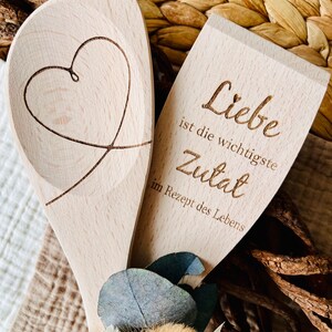 May include: Two wooden spoons, one with a heart carved into it and the other with the text "Liebe ist die wichtigste Zutat im Rezept des Lebens" (Love is the most important ingredient in the recipe of life).