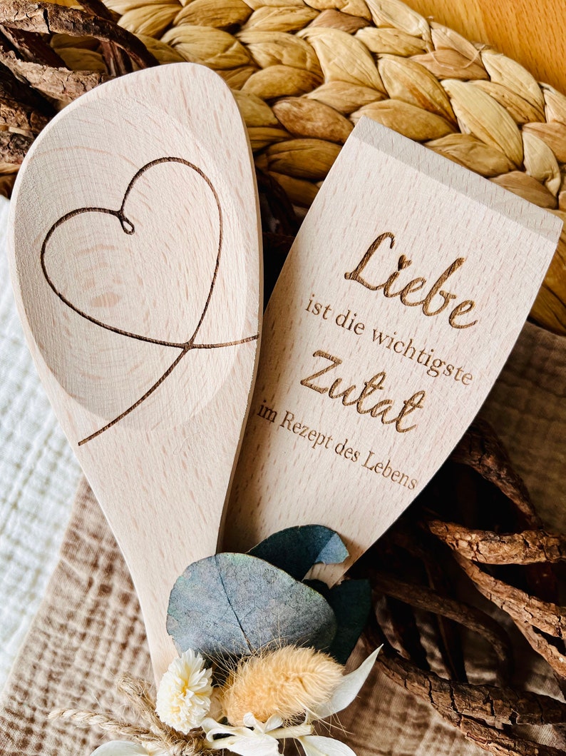 May include: Two wooden spoons, one with a heart carved into it and the other with the text "Liebe ist die wichtigste Zutat im Rezept des Lebens" (Love is the most important ingredient in the recipe of life).