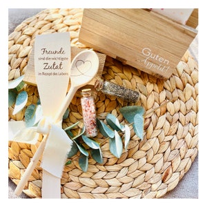 May include: Wooden spoon with the text "Friends are the most important ingredient in the recipe of life" and a heart carved into the spoon. Two small glass jars with spices are on a woven placemat with a wooden box that says "Guten Appetit!"