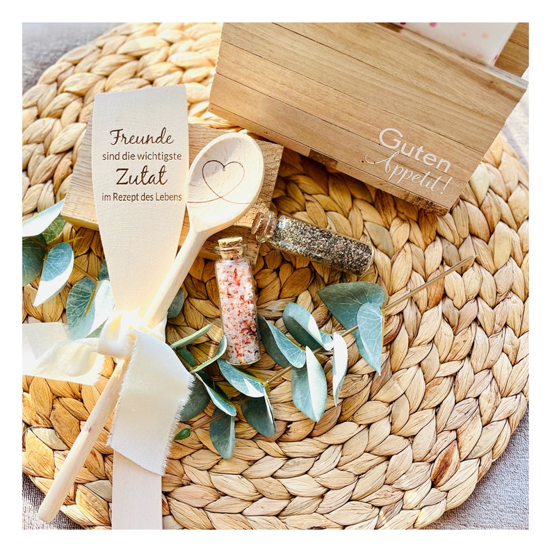 May include: Wooden spoon with the text "Friends are the most important ingredient in the recipe of life" and a heart carved into the spoon. Two small glass jars with spices are on a woven placemat with a wooden box that says "Guten Appetit!"