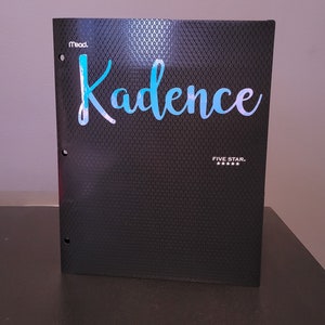 Könnte beinhalten: Ein schwarzer Ordner mit einer strukturierten Oberfläche. Der Ordner hat drei Löcher auf der linken Seite und den Text "Mead" und "Five Star" aufgedruckt. Der Name "Kadence" ist in blauer Kursivschrift auf der Vorderseite des Ordners geschrieben.