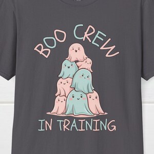 May include: Dark grey t-shirt with the text "BOO CREW IN TRAINING" in pink and blue. The shirt features a stack of cartoon ghosts in pink and blue with smiling faces. The shirt is made of a soft material.