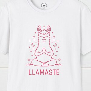 Op de afbeelding: Wit T-shirt met een roze omtrek van een mediterende lama met de tekst "LLAMASTE" eronder.