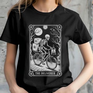 Puede incluir: Camiseta negra con un diseño gráfico en blanco y negro de un esqueleto en bicicleta, entregando paquetes. El diseño incluye una luna llena, un cuervo y el texto "THE DELIVERER". La camiseta es de cuello redondo.