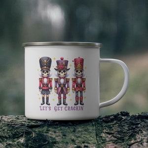 May include: White enamel mug featuring three skeletal nutcracker figures in red, purple, and black uniforms. The mug has a silver rim and a white handle. The text "LET'S GET CRACKIN'" is printed below the figures.