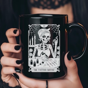 May include: Black ceramic mug featuring a white illustration of a skeleton tattoo artist. The design includes candles, spiderwebs, and the text "THE TATTOO ARTIST". The mug has a large handle.