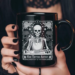 May include: Black ceramic mug featuring a skeleton tattoo artist design. The design includes a skeleton holding a tattoo gun, surrounded by roses and skulls, with the text "The Tattoo Artist".