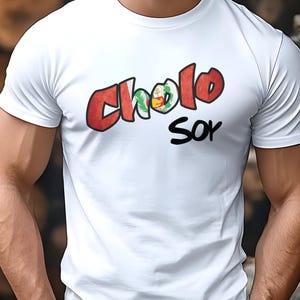 Op de afbeelding: Wit T-shirt met de tekst "Cholo Soy" in rode en zwarte letters. Het ontwerp bevat een klein embleem met groene, gele en rode kleuren. Het shirt met korte mouwen lijkt van katoen te zijn.