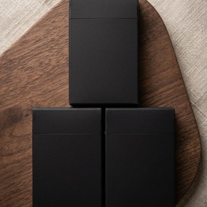 May include: Three black rectangular boxes with a matte finish are arranged on a wooden cutting board. The boxes are evenly spaced and have a simple, minimalist design. The background is a neutral-colored fabric.