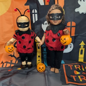 May include: Two dolls dressed in red ladybug costumes with black polka dots, black pants, and black glittery eye masks. The smaller doll wears a ladybug headband with antennae. Both dolls hold small orange plastic jack-o'-lantern candy buckets.