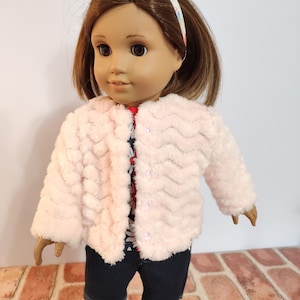 May include: A pink faux fur jacket with a button closure, worn by a doll with brown hair and a floral print shirt. The doll is wearing blue denim shorts and yellow shoes.