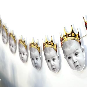 May include: A garland of baby faces with gold crowns. The black and white images of the babies are strung together with twine. The crowns are gold with red and black accents. The garland is hanging against a white wall.