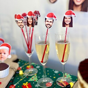 Op de afbeelding: Een feestelijke kerstparty-scène met twee glazen champagne, elk met een rode rietje en een feestelijke kerstmanmuts met een gezicht erop. De glazen staan op een groene tafelkleed met een kerstpatroon. Er zijn andere kerstmanmützen met gezichten erop op de achtergrond.