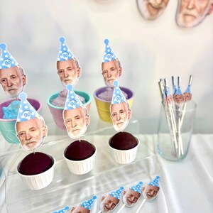 May include: Cupcakes with dark frosting and decorative toppers featuring a person's face wearing a blue party hat with the number 90. The cupcakes are in white paper cups and pastel-colored containers.