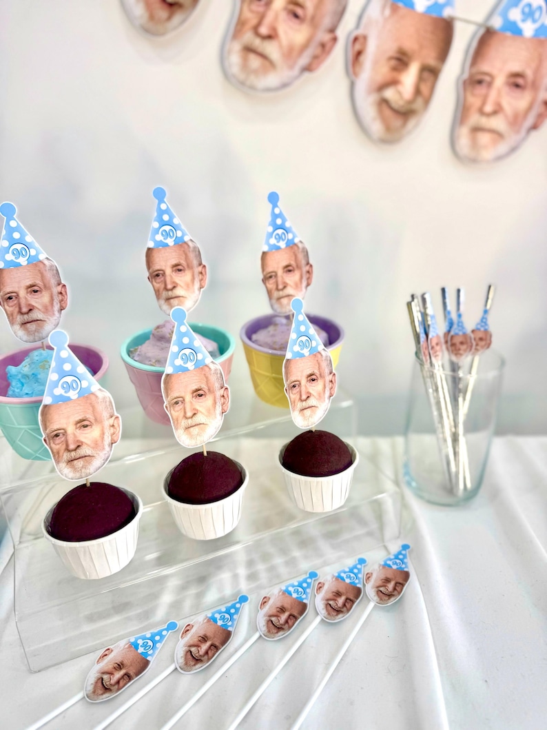 May include: Cupcakes with dark frosting and decorative toppers featuring a person's face wearing a blue party hat with the number 90. The cupcakes are in white paper cups and pastel-colored containers.