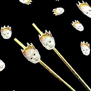 Op de afbeelding: Een zwarte achtergrond met verspreide babygezichten met gouden kronen. Twee goudkleurige stokken zijn bekroond met babygezichten met gouden kronen. De babygezichten zijn wit met een neutrale uitdrukking.