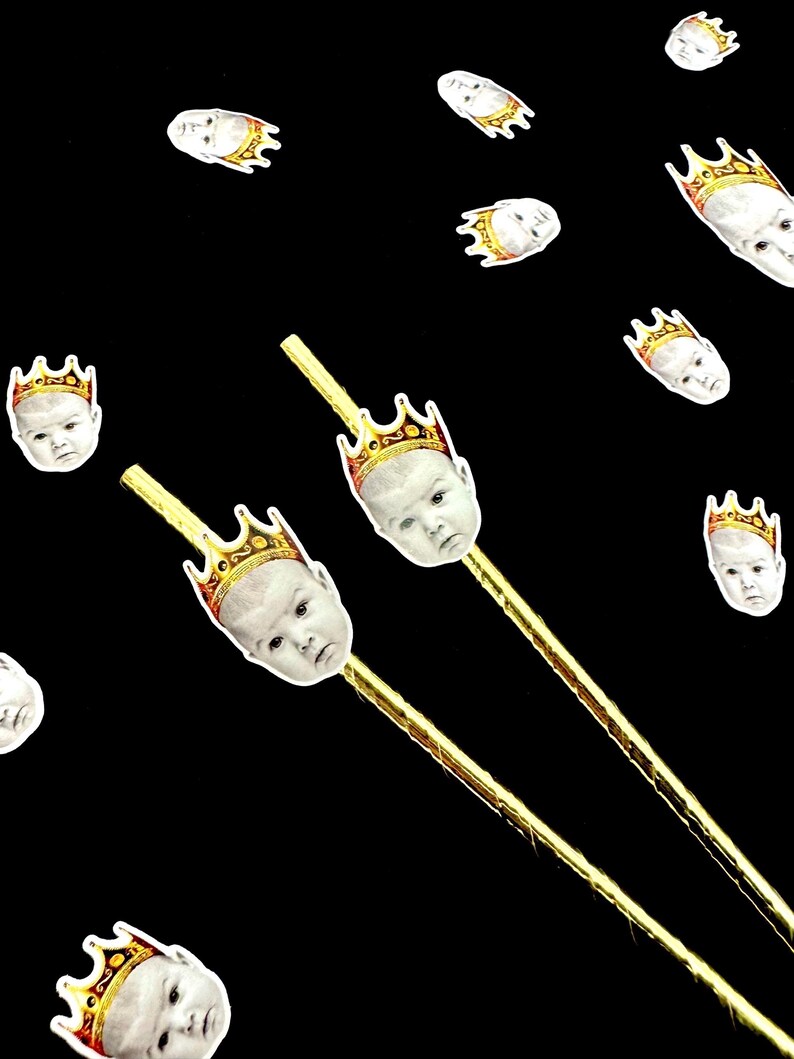 Op de afbeelding: Een zwarte achtergrond met verspreide babygezichten met gouden kronen. Twee goudkleurige stokken zijn bekroond met babygezichten met gouden kronen. De babygezichten zijn wit met een neutrale uitdrukking.