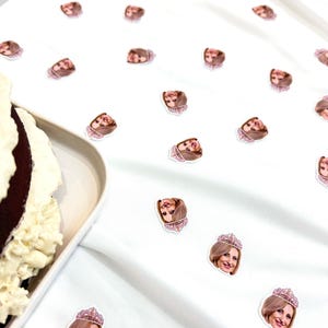 May include: White tablecloth scattered with small stickers featuring a woman's face wearing a pink jeweled crown. A partially visible chocolate cake with white frosting sits on a white plate.