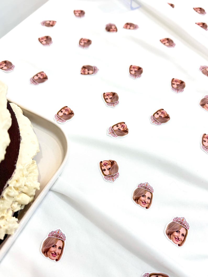 May include: White tablecloth scattered with small stickers featuring a woman's face wearing a pink jeweled crown. A partially visible chocolate cake with white frosting sits on a white plate.