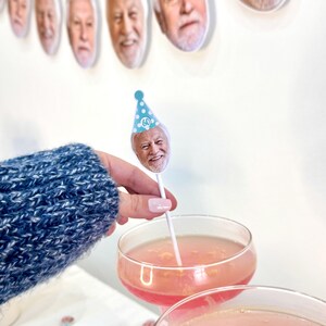 K&ouml;nnte beinhalten: Eine festliche Geburtstagsszene mit einem Banner aus l&auml;chelnden Gesichtern mit Partyh&uuml;ten. Ein Cocktail ist mit einem Gesicht auf einem Stiel garniert, das einen Partyhut mit der Nummer 60 tr&auml;gt. Das Getr&auml;nk ist rosa und wird in einem Coup&eacute;-Glas serviert.