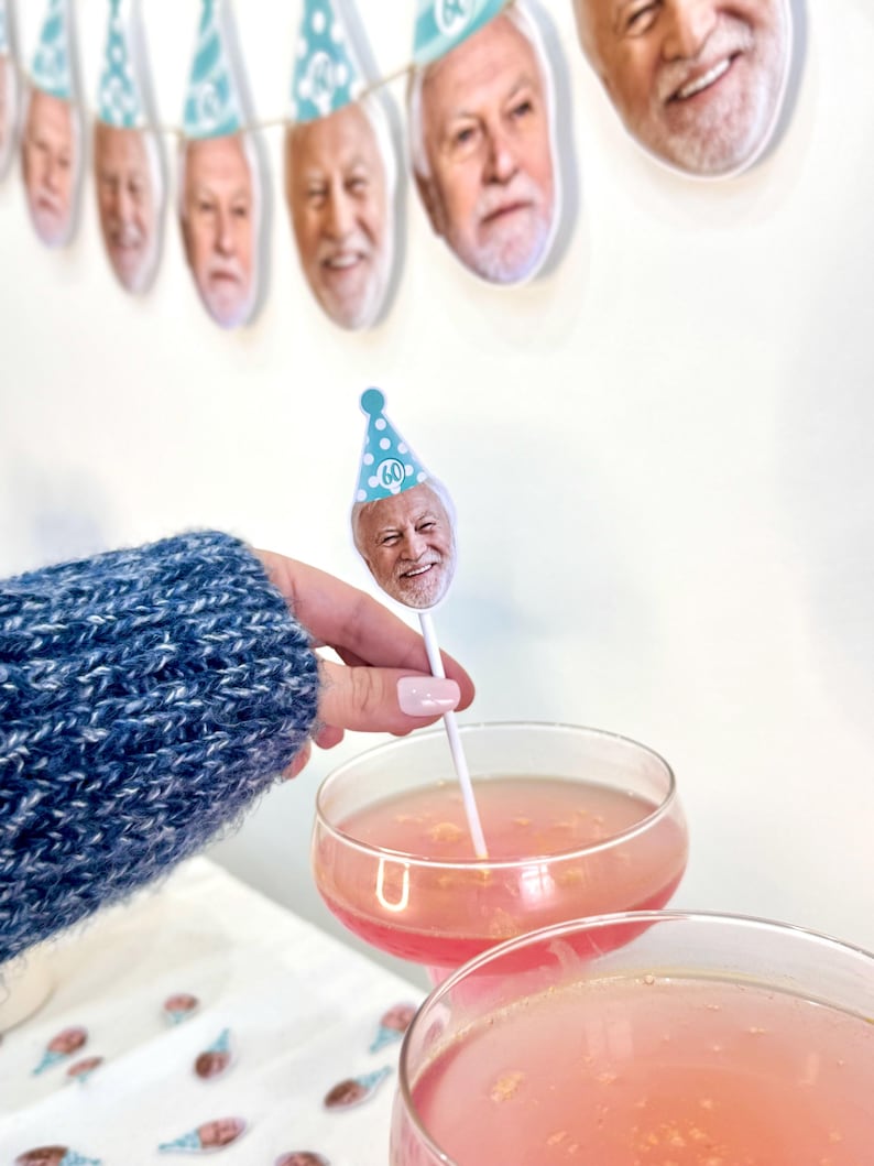 K&ouml;nnte beinhalten: Eine festliche Geburtstagsszene mit einem Banner aus l&auml;chelnden Gesichtern mit Partyh&uuml;ten. Ein Cocktail ist mit einem Gesicht auf einem Stiel garniert, das einen Partyhut mit der Nummer 60 tr&auml;gt. Das Getr&auml;nk ist rosa und wird in einem Coup&eacute;-Glas serviert.