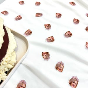 May include: White fabric scattered with small stickers featuring a woman's face wearing a tiara. A partially visible cake with white frosting and a brown base sits on a white plate to the left.
