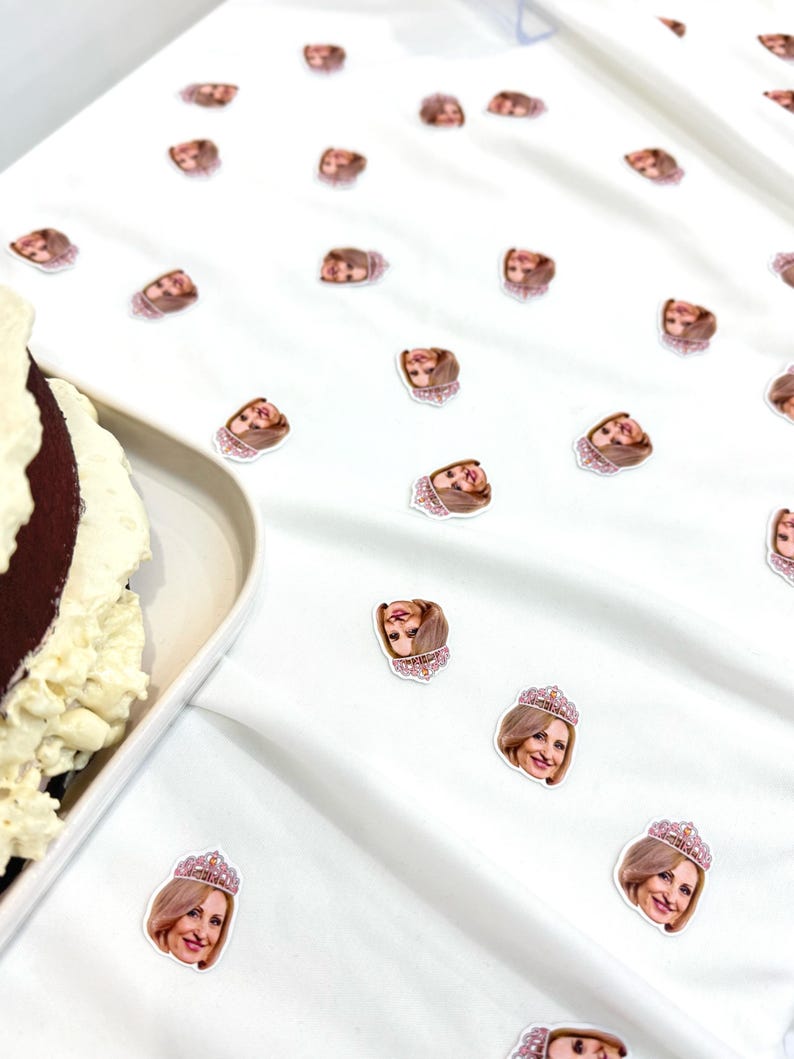 May include: White fabric scattered with small stickers featuring a woman's face wearing a tiara. A partially visible cake with white frosting and a brown base sits on a white plate to the left.
