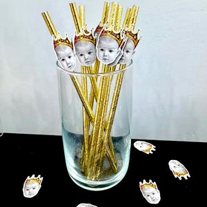 Op de afbeelding: Een helder glas gevuld met goudkleurige rietjes, elk versierd met babygezichten met kronen. Verschillende babygezicht-uitsparingen met kronen zijn verspreid over het zwarte oppervlak. De rietjes zijn gerangschikt in een helder glas.