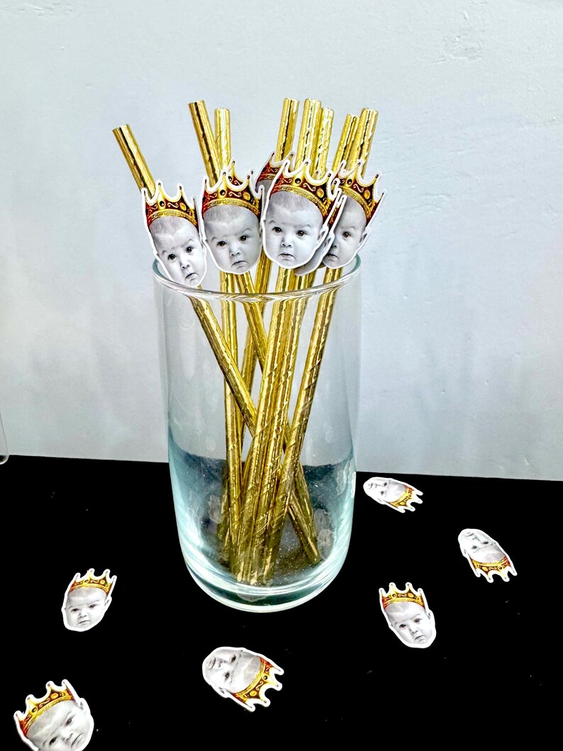 Op de afbeelding: Een helder glas gevuld met goudkleurige rietjes, elk versierd met babygezichten met kronen. Verschillende babygezicht-uitsparingen met kronen zijn verspreid over het zwarte oppervlak. De rietjes zijn gerangschikt in een helder glas.