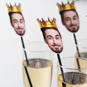K&ouml;nnte beinhalten: Champagnergl&auml;ser mit einem Foto-Requisit eines Mannes mit einer goldenen Krone. Die Requisiten befinden sich auf schwarzen St&auml;ben. Der Champagner hat eine hellgelbe Farbe.