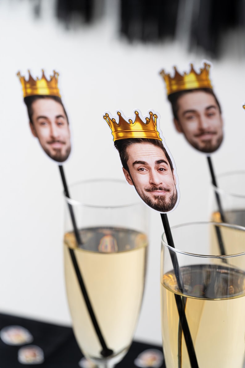 K&ouml;nnte beinhalten: Champagnergl&auml;ser mit einem Foto-Requisit eines Mannes mit einer goldenen Krone. Die Requisiten befinden sich auf schwarzen St&auml;ben. Der Champagner hat eine hellgelbe Farbe.