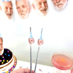 May include: Two silver straws topped with a cutout of a man's face wearing a blue party hat. A banner of similar faces in party hats hangs in the background. A frosted cake and pink drink are also visible.