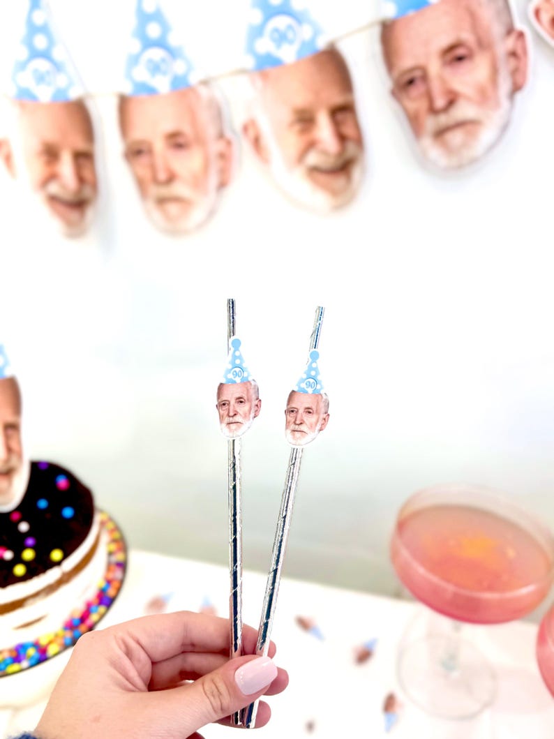 May include: Two silver straws topped with a cutout of a man's face wearing a blue party hat. A banner of similar faces in party hats hangs in the background. A frosted cake and pink drink are also visible.