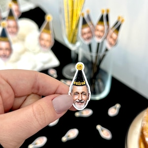 May include: A close-up of a small, clear sticker with a smiling face wearing a black and gold party hat with the word "retired" printed on it. The sticker is held by a hand. Other stickers and cocktail stirrers with the same face are in the background.