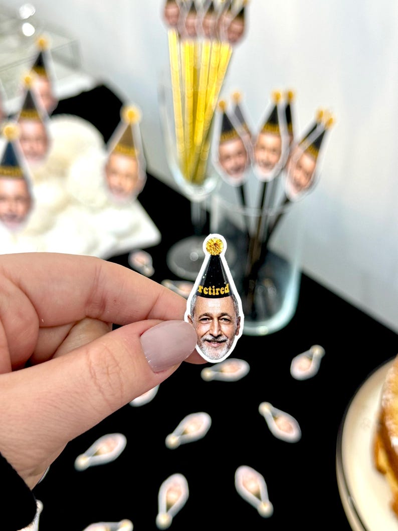 May include: A close-up of a small, clear sticker with a smiling face wearing a black and gold party hat with the word "retired" printed on it. The sticker is held by a hand. Other stickers and cocktail stirrers with the same face are in the background.