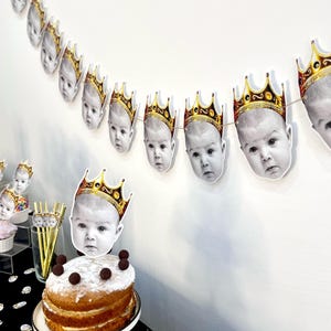 May include: A festive party scene featuring a cake and garland. The cake is topped with a baby's face wearing a gold crown. A garland of baby faces with gold crowns hangs above. The cake is on a white cake stand, and the table is covered with a black tablecloth.